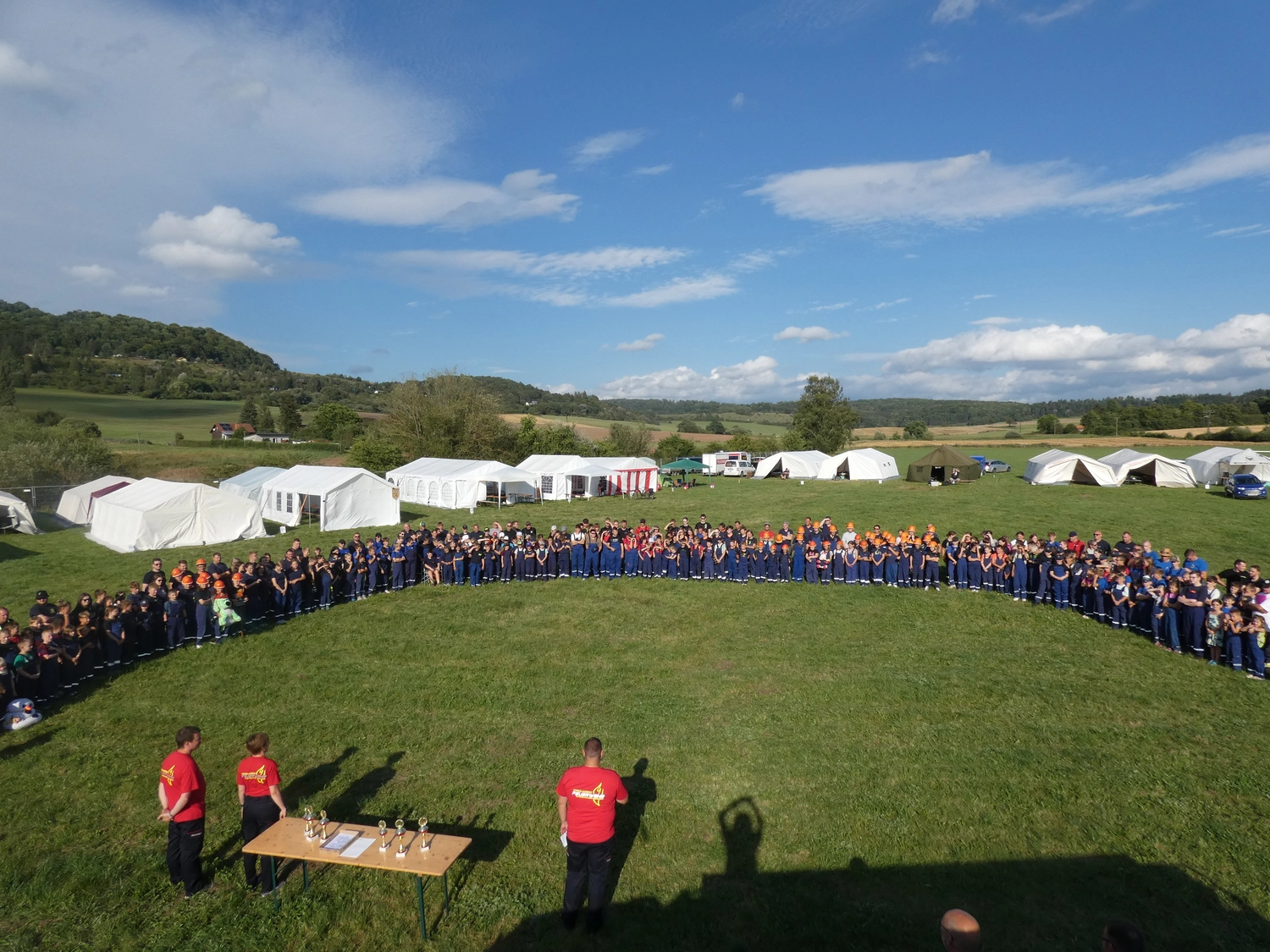 Krerisfeuerwehrverband Schmalkalden-Meiningen e.V. - Koordiniert die Aktivitäten und Veranstaltungen der Feuerwehr - Eine große Gruppe von Menschen in blauen Uniformen bildet einen weiten Halbkreis auf einem grasbewachsenen Feld; im Hintergrund sind weiße Zelte und sanfte Hügel unter blauem Himmel zu sehen.