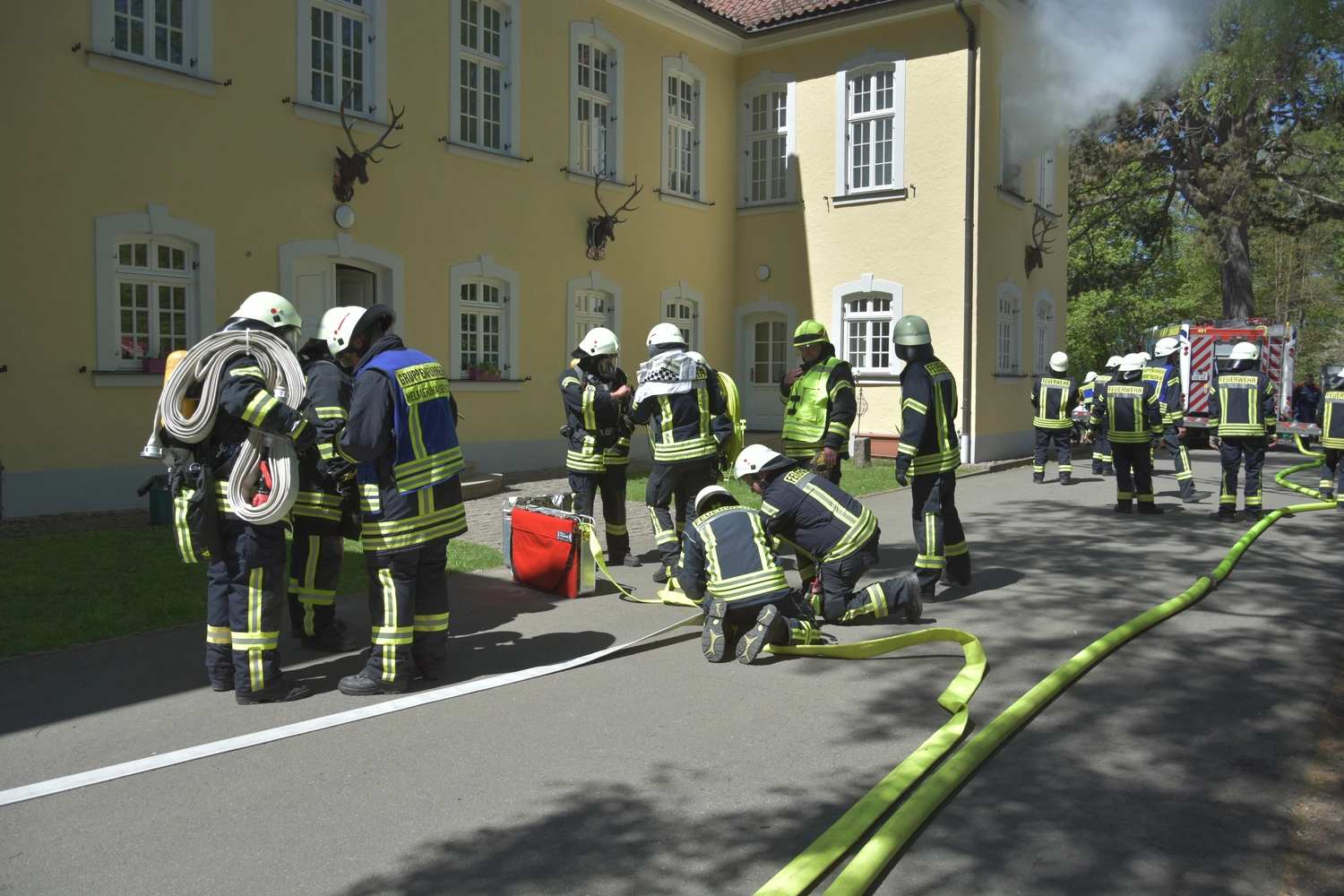 Krerisfeuerwehrverband Schmalkalden-Meiningen e.V. - Koordiniert die Aktivitäten und Veranstaltungen der Feuerwehr - Feuerwehrleute in orangefarbenen Helmen und blauen Uniformen nehmen an einer Übung teil, bei der sie mit einem Schlauch einen Brand in einem kleinen Holzgebäude löschen, während ein anderer Auszubildender kniet und zusieht.